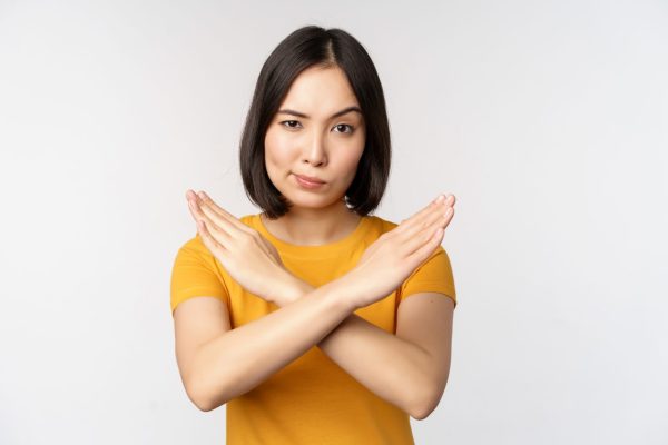 Portrait of asian woman looking serious and angry, showing stop prohibit gesture, taboo sign, forbidding smth, standing in yellow tshirt over white background.